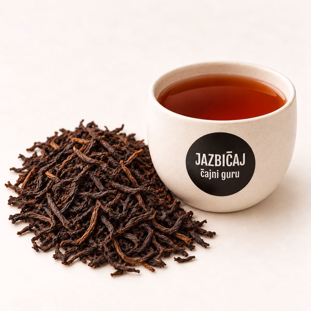 Skodelica črnega čaja z logotipom Jazbičaj ob kupčku posušenih listov črnega čaja.