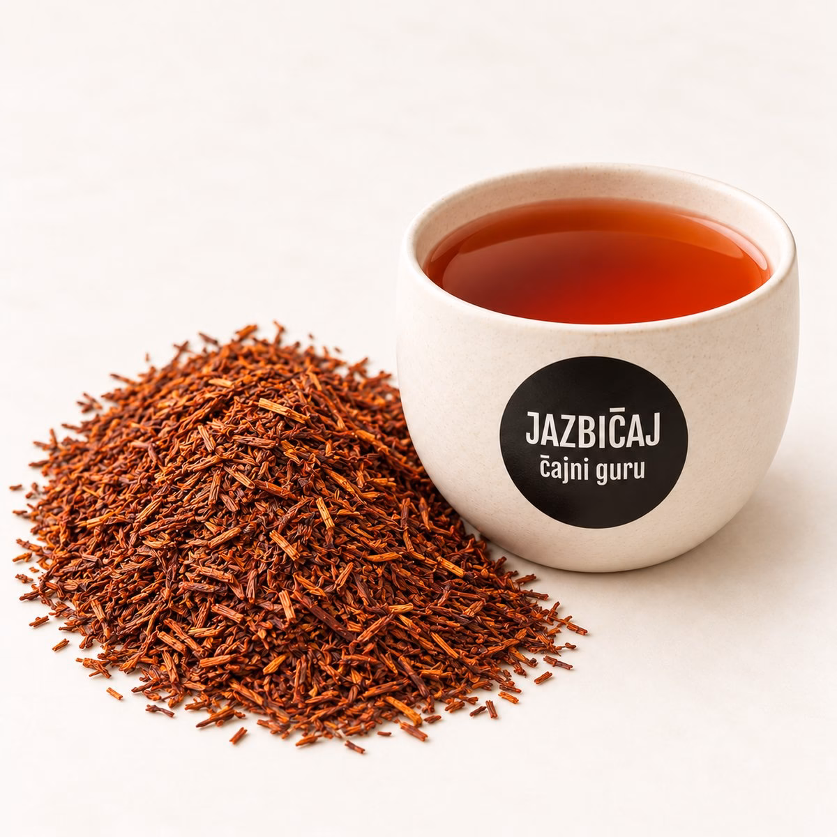 Skodelica rooibos čaja z logotipom Jazbičaj ob kupčku drobnih posušenih lističev rooibosa.
